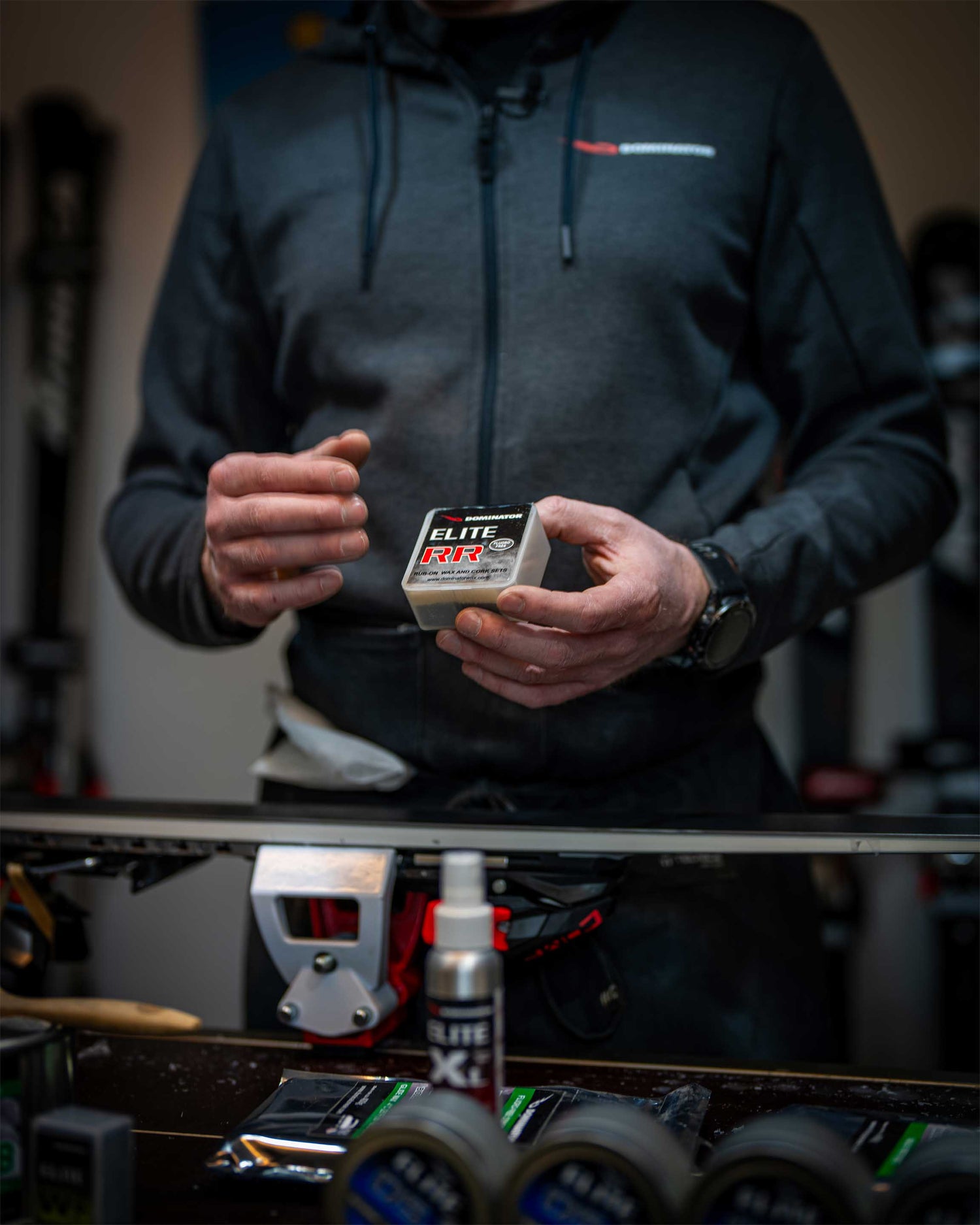 Ein Dominator Wax Elite RR wird in einer Skiwerkstatt in der Hand gehalten, während ein Ski auf einer Arbeitsbank liegt. Im Vordergrund sind weitere verschwommene Dominator-Produkte sichtbar. Das Bild unterstreicht die Verbindung von Technologie und Handwerkskunst.