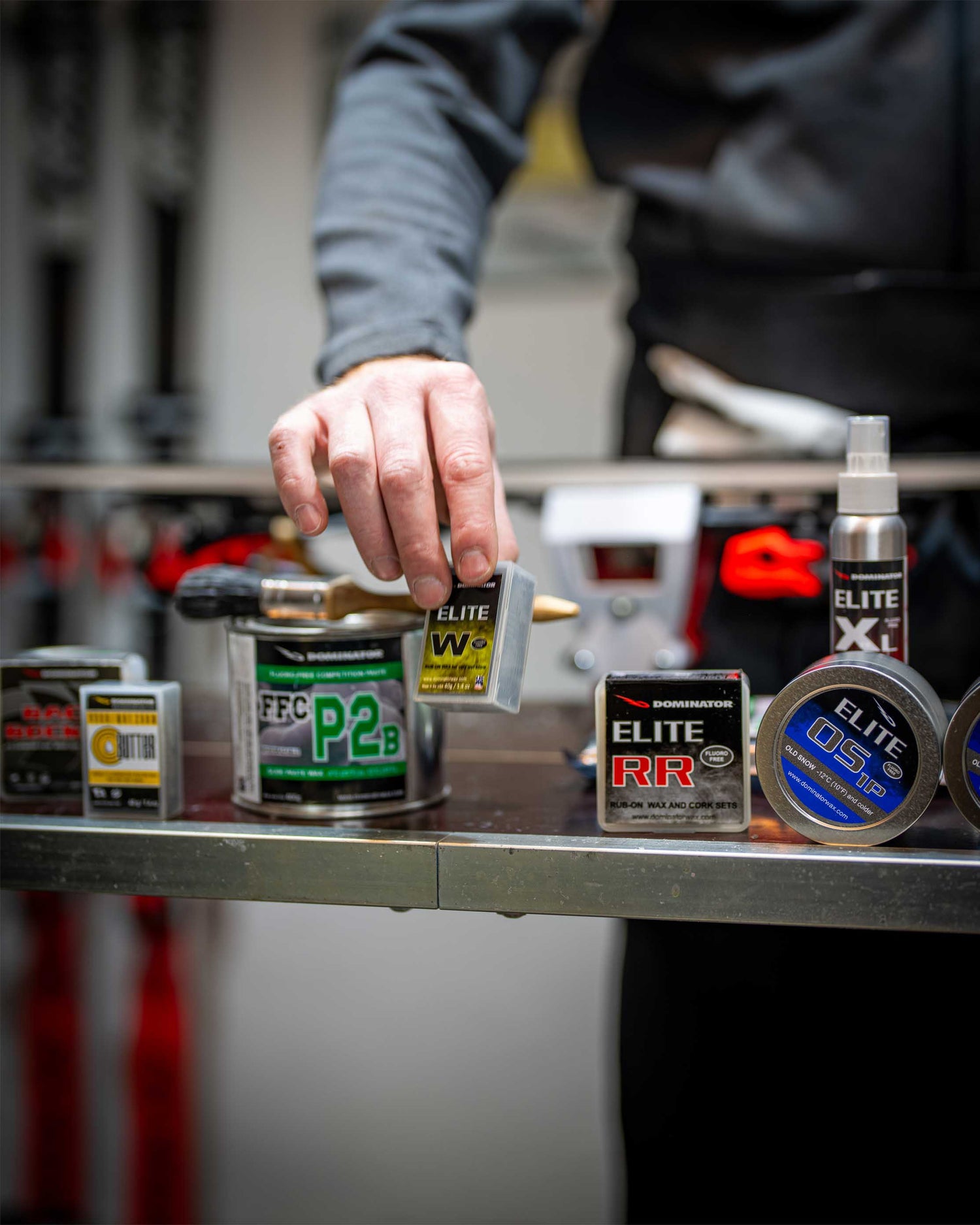 Nahaufnahme eines Dominator Elite W Produkts, das in einer Skiwerkstatt aus einem Regal genommen wird. Im Hintergrund sind weitere Dominator-Produkte und ein Ski sichtbar, die die Vielfalt und Qualität des Angebots zeigen.