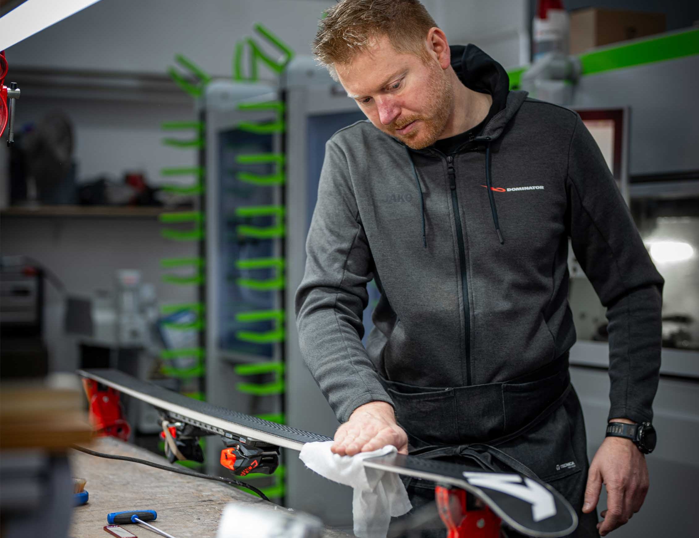 Eine Person bereitet einen Ski mit einem Lappen für das Wachsen vor. Der Ski liegt auf einer Arbeitsplatte, und die Szene betont die professionelle Pflege und das Engagement der Dominator-Partnerhändler.