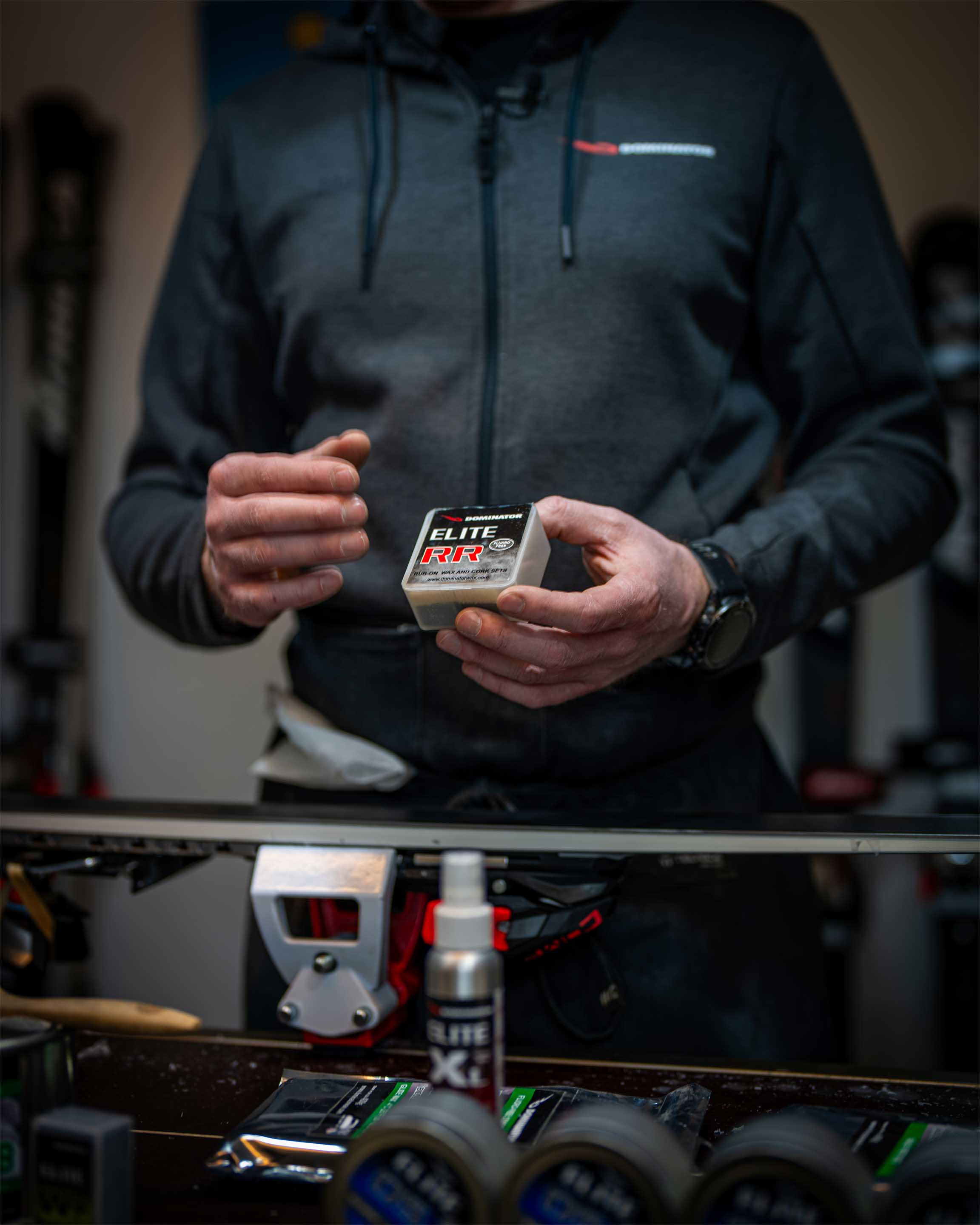 Ein Dominator Wax Elite RR wird in einer Skiwerkstatt in der Hand gehalten, während ein Ski auf einer Arbeitsbank liegt. Im Vordergrund sind weitere verschwommene Dominator-Produkte sichtbar. Das Bild unterstreicht die Verbindung von Technologie und Handwerkskunst.